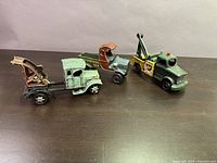 Three vintage die cast toy trucks displayed on a dark surface: a light blue tow truck, a green and red truck with an open cargo bed, and a green tow truck with a crane.