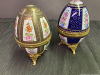 Two Limoges porcelain egg-shaped trinket boxes with floral decoration and gold-tone accents on ornate feet.