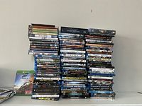 Photo showing a large collection of DVDs and Blu-ray movies stacked in multiple columns, showing a variety of movie titles and Xbox games, some unopened.