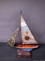 Metal sailboat sculpture with stylized map and compass rose on main sail, green swirled small sail, wooden hull, and bird on mast, viewed frontally.