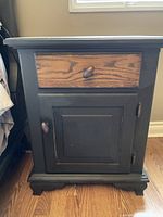 Front view of the night table highlighting the black-painted frame, natural wood drawer front, door with knob, and bracket feet.