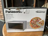 Exterior of boxed Panasonic microwave (model NN-SC73LS) showing brand logo, inverter label, and pizza graphic.