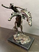 Front angled view of bronze cowboy on horse sculpture showing the cowboy with one arm raised and the horse bucking.