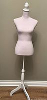 Full view of sewing and staging manikin with linen torso and wooden tripod stand against tan wall.