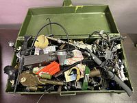 Green military-style plastic case filled with assorted GI Joe accessories including rifles, helmets, scopes, and other gear.