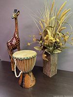 Photo showing the giraffe statue, wooden drum, and floral arrangement together on a table against a plain background.