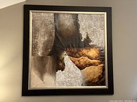 Front-facing image of the large square abstract painting hanging on a wall, showing textures and colors in brown, gold, gray, and black, framed with a wide black and gold edged frame.