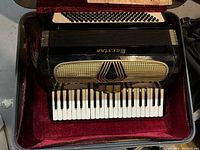 Top-down photo of a vintage black Masola accordion with standard piano keyboard and multiple bass buttons in a red velvet-lined case.