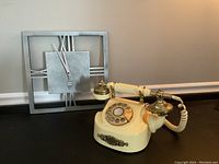 Photo showing a stainless steel wall clock with open face design and a cream vintage rotary dial telephone with brass details on a flat surface.