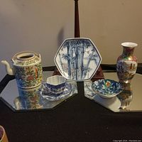 Wide view showing entire set of five Asian ceramic pieces on mirrored surfaces: teapot, cup with saucer, hexagonal plate, bowl, and vase.