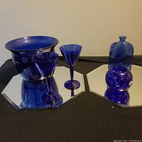 Full view of five cobalt blue glass items on mirrored platforms including bowl, goblets, bottle, and vase.
