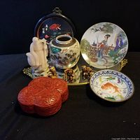 Overview of the entire lot displayed on a gold trimmed ornate mirror tray showing the porcelain vase, large and small porcelain plates, three carved figurines, red carved lacquer box, black lacquer plaque with floral inlay.