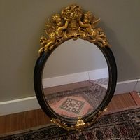 Full view of the oval decorative wall mirror showing the ornate black and gold frame with cherubs and floral design.