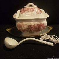 Full view of electric soup tureen urn and ceramic ladle with cord on a black background, showing red scenic transferware pattern and lid.