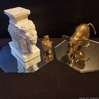 Three elephant figurines: ivory-colored ornate elephant, small gold-tone elephant, large brass elephant on mirror-like surface.