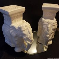 Pair of white ceramic elephant stands shown side by side on a reflective surface with detailed elephant heads and embossed tops visible