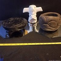 Photo showing the three items including the black carved wooden stool, white ceramic bird figurine, and carved wooden vessel, placed on mirror bases with a measuring tape in front.