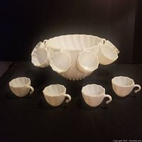 Full set including one large punch bowl and eight matching milk glass cups with handles arranged around it on a black background.