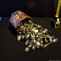 Photograph showing a large quantity of vintage collector spoons spilling from a colorful vintage-style tin container.