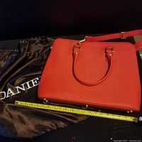 Full view of red leather handbag with dust cover and measuring tape showing width.
