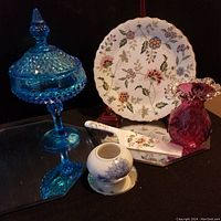 Photo showing blue press glass lidded pedestal candy dish, pink ruffled glass vase, floral pie plate, ceramic cake server, and porcelain jar.