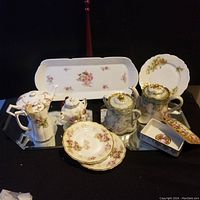 Overview of the collection showing multiple porcelain items on reflective surface including trays, teapots, plates, and condiment dishes all with floral and gold trim decoration