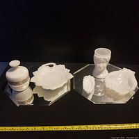 Photo showing six vintage milk glass items arranged on an octagonal mirrored surface. Items include a lidded jar, scalloped bowl, head-profile container, and three hand-shaped dishes in various sizes.