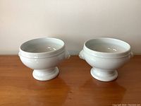 Pair of white ceramic vessels with pedestal bases and lion head design on sides, shown from front angle.