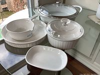 Full view of all cookware items set on glass table including large oval casserole with glass lid, medium round casserole with lid, divided bowl, rectangular dish, and plate