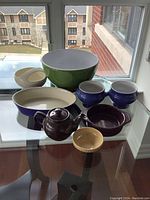 Photo showing the full set of assorted colored dishes including large green bowl, teapot, various bowls, and an oval serving dish on glass-top table.