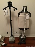 Full view of two garment steamers side by side showing bases, poles, hangers, hoses, and instruction tags.