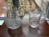 Both water pitchers side by side on a wooden surface by a window, viewed frontally showing cut patterns and handles.