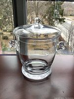 Front view of a clear hand blown glass ice bucket with a rounded lid and bulbous knob handle on top, placed on a wooden surface with a window background.