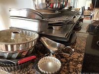 Wide shot showing metal baking trays stacked, bundt cake pan, muffin tins, tart pans, metal funnel, and other baking accessories on kitchen counter