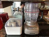 Full view of the KitchenAid Architect food processor with accessories in a white storage container beside it, placed on a kitchen counter.