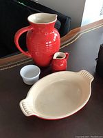 All four pieces of Le Creuset kitchenware displayed on table: large red pitcher, small red creamer, white ramekin, and oval baking dish