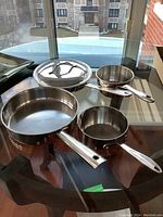 Overall view of all four cookware items placed on a glass table including large skillet, two saucepans, and a lid