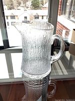 Clear textured glass pitcher on reflective glass table near window, showing whole pitcher with handle and spout from the side.