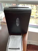 Front and top view of black Staples paper shredder with visible crack on top, sitting near window, including operator's manual in front.