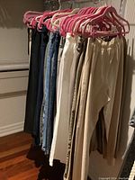 Photo shows a variety of women's pants hanging on rod with pink and white hangers, featuring multiple colors and styles including denim, neutral tone pants with elastic waistbands.