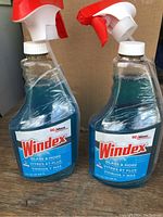 Two Windex Glass & More spray bottles placed on wooden surface with cardboard background, showing blue cleaner liquid and white-red spray nozzles