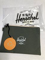 Photo showing olive green Herschel pouch with front logo and attached tag, next to clear plastic Herschel branded packaging.