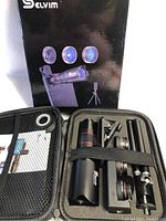 Opened carrying case showing a telephoto lens, two smaller lenses, clip for attaching lenses to phone, and small tripod secured by elastic bands in the case.