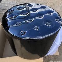 Photo showing round black ottoman with shiny tufted top surface and smooth sides, positioned partly on white protective wrapping and cardboard.