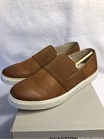 Side view of the left and right cognac Kenneth Cole loafers on white box, showing the shoe profile and upper ribbed elastic strap