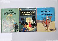 Photo of three softcover Tin Tin comic books laid flat showing front covers: The Seven Crystal Balls, The Castafiore Emerald, The Crab with the Golden Claws.
