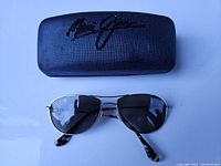 Sunglasses and hard case overview showing sunglasses with silver metal frame and dark lenses below a black textured case with large Maui Jim signature on top.