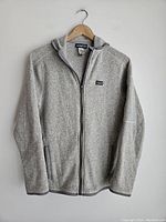 Front view of light grey Patagonia fleece jacket with hood hanging on wooden hanger showing full zip and Patagonia logo patch on chest.