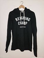 Front view of black Reigning Champ hoodie hanging on wooden hanger, showing white printed brand name and athletic gear text on chest.