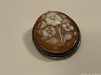 Close-up front view of the oval floral cameo brooch showing detailed white flower carvings on brown background and silver twisted wire frame.